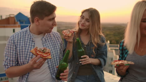 Loving-couple-on-the-roof-clinks-beer-from-green-bottles-and-eats-pizza.-Girl-shyly-and-tenderly-looks-at-her-boyfriend.